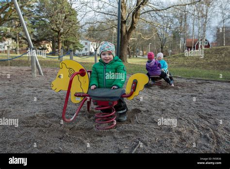 Symbolbild: Eine Mutter mit ihrem ersten Kind auf einem Spielplatz, die nachdenklich auf eine Familie mit zwei Kindern blickt.